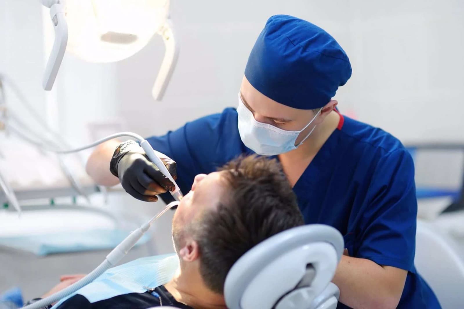 A dentist in blue scrubs and a surgical cap treating a patient in the dental chair